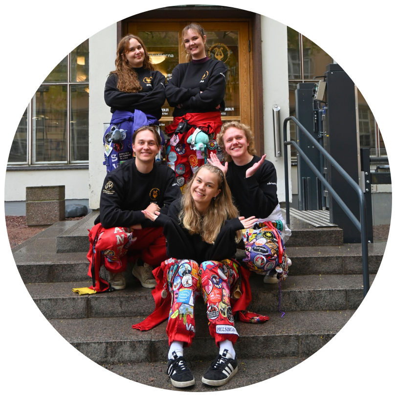 ÅAS styrelse 2026 står och sitter på trappor och ler. Alla har halare på sig och svarta tröjor med ÅAS logo på brösten.