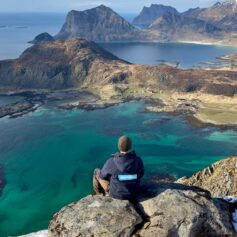 En människa sitter ryggen mot kameran på en klippa på Lofoten. På bild syns klart blått vatten och klippor.