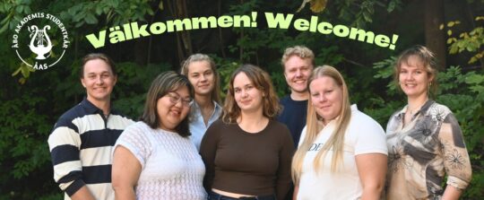 ÅAS styrelse 2026. Sju människor står ute, tittar mot kameran och ler. Det finns träd och gröna blad i bakgrunden och en text Välkommen Welcome ovanför människorna.