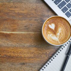 Foto på en kaffedryck på ett bord, i hörnet syns en bit av en bärbar dator samt anteckningsblock och penna